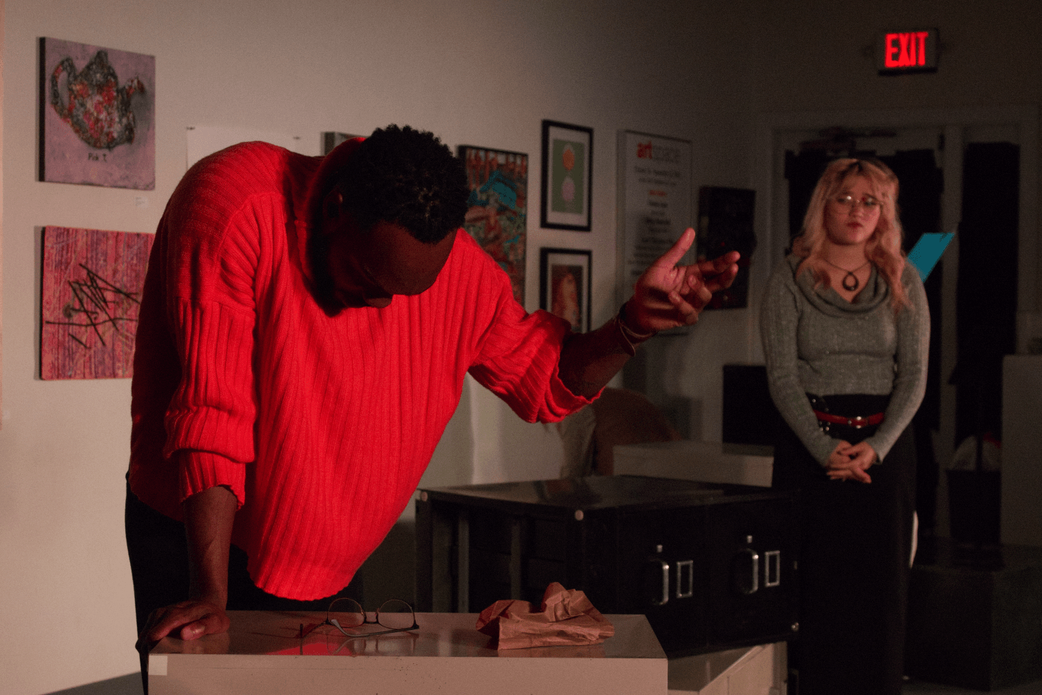 a character in a bright orange sweater with his head down towards a file cabinet and one arm raised